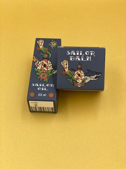 Two navy printed boxes, a tall narrow one for a 30 ml oil and a small square one for a balm, with a nautical tattoo-style illustration